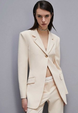 Blazer crema con solapas de pico, un botón y bolsillos frontales. Combinado con pantalones crema a juego, de tejido suave y corte entallado.