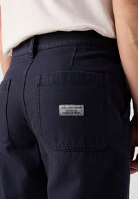 Tamno plave traperice glatke teksture, s stražnjim džepom i detaljem ušivenom oznakom blizu džepa.