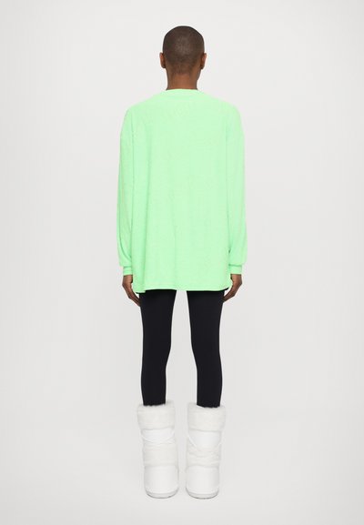 Maglietta a maniche lunghe verde neon con collo rotondo, abbinata a leggings neri e stivali foderati in pelliccia bianca, vista dal retro.