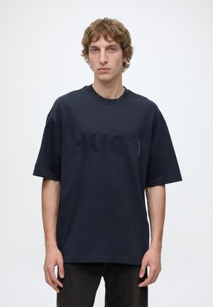 Jeune homme aux cheveux bouclés portant un t-shirt ample bleu marine foncé avec un texte en relief "HUGO", debout devant un fond clair uni.