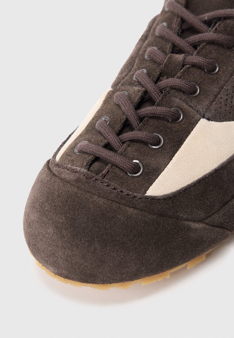 Sneaker in suede marrone con lacci neri, dettagli beige e punta arrotondata con suola in gomma testurizzata. Perforazioni sottili sulla parte superiore.