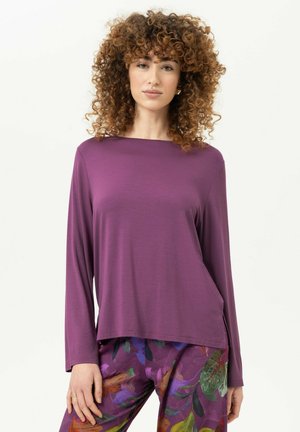 Frau mit lockigem Haar, die ein langärmeliges violettes Oberteil und geblümte Hose trägt, steht vor einem einfarbigen hellen Hintergrund.