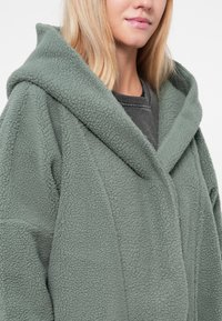 Grünfleece-Hoodie mit weicher Textur, Vordertaschen und lässiger Silhouette, ausgestattet mit einem breiten, umgeschlagenen Kragen.