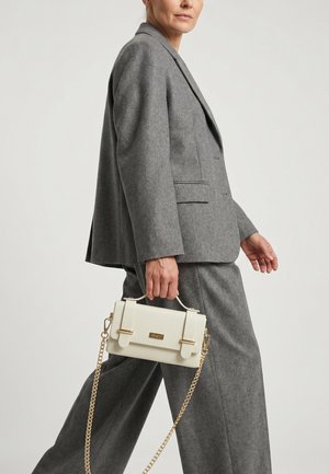 Žena v šedém kostýmku drží malou bílou kabelku se zlatým řetízkem a kovovými doplňky, kráčející proti jednobarevnému pozadí.