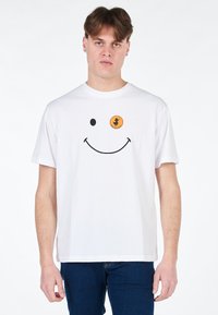 T-shirt bianca in cotone con una grafica di faccina sorridente nera e un'etichetta circolare arancione con testo, il tutto su uno sfondo semplice.