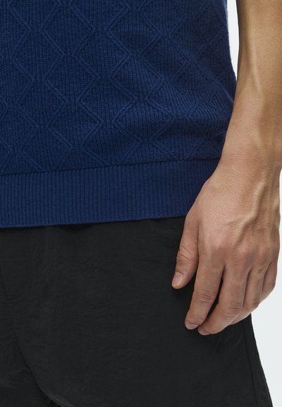Pull en maille bleu marine présentant un motif en losange texturé, avec un ourlet côtelé. La main visible repose contre un tissu noir texturé.