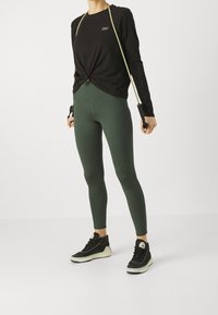 Svart långärmad tröja med en vriden knutdesign, gröna leggings och svarta sneakers med vit sula. Slätt tyg och åtsittande stil.
