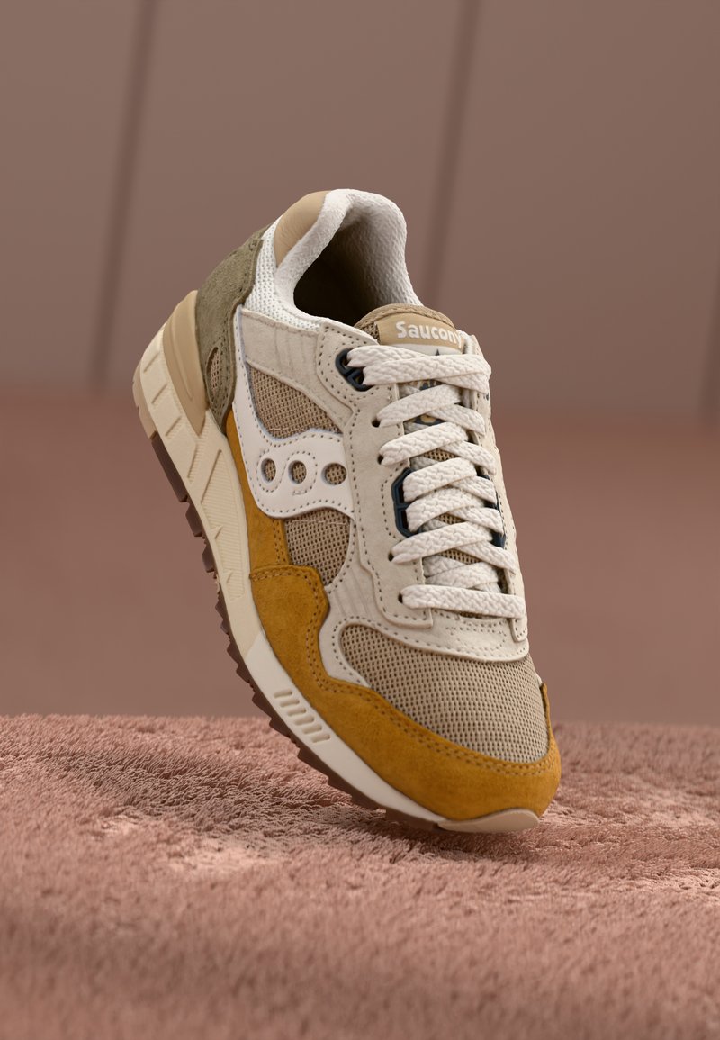 Beige and yellow sneaker with textured suede and mesh panels, white laces, and Saucony branding on the side, set on a soft pink surface.