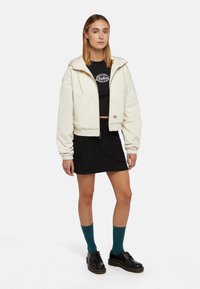 Chaqueta corta de color blanco roto con capucha, cierre de cremallera y puños acanalados, combinada con un top corto negro, una falda negra, calcetines en color teal y zapatos negros.