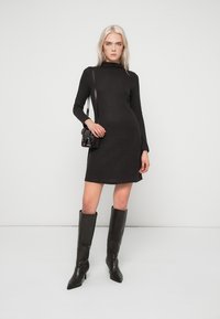 Vestido de malha preto com gola alta, mangas compridas e bainha ligeiramente evasê. Combinado com botas pretas em pele até ao joelho e uma pequena mala.