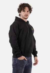 Felpa nera con tasca a marsupio frontale, cappuccio con coulisse e un piccolo logo verde sul petto. Realizzata in un morbido tessuto testurizzato.