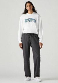 Camicia bianca a maniche lunghe con logo "Levi's" colorato, abbinata a pantaloni grigi scuri con vita elastica e sneaker bianche. Design semplice, vestibilità casual.
