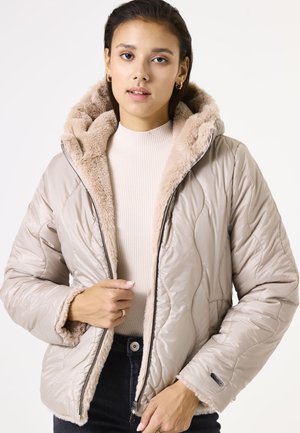 Beige gewatteerde jas met een nepbont capuchon en binnenvoering. Voorzien van een ritsluiting en een textuurstof. Gepaard met een licht geribbeld topje.