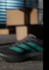 Black sneaker with teal stripes on a wooden floor, blurred background with stacked cardboard boxes and window blinds.