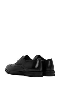 Par de zapatos de vestir de cuero negro tipo brogue con perforaciones decorativas y tacones bajos apilados, mostrados desde el ángulo trasero y lateral.