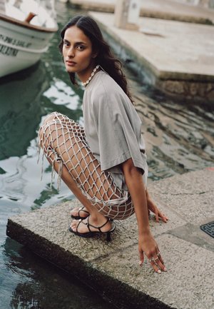 Hosszú sötét hajú nő bézs felsőben és hálós szoknyában guggol a csatorna szélén, fekete pántos magassarkú cipőben és kagyló nyaklánccal.