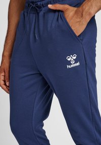 Mörkblå mjukisbyxor med dragsko i midjan, sidofickor och en vit "hummel"-logotyp tryckt på låret. Slät, mjuk tygtextur.