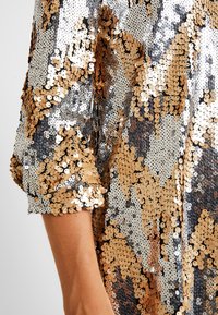 Top sekaný ve zlaté a stříbrné barvě s geometrickým vzorem. Má volný střih a rukávy s manžetami. Texturovaný povrch díky překrývajícím se flitrům.