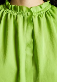 Grüne Bluse mit Rüsche am Kragen, die eine glatte Textur und einen dezenten Glanz aufweist. Der Stoff hat einen leichten Fall, der das Gesamtdesign unterstreicht.