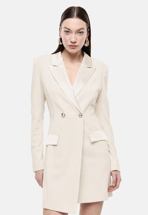 Donna con capelli lisci e scuri indossa un abito blazer doppiopetto color crema con spalle imbottite e grandi orecchini a cerchio dorati.