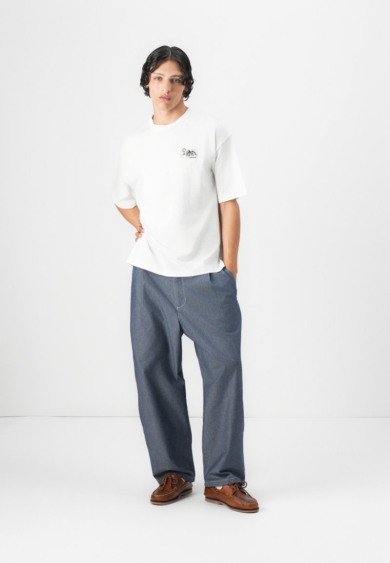 T-shirt blanche à manches courtes avec un petit graphique noir, associée à un pantalon en denim bleu ample et des chaussures en cuir marron à lacets.