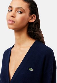 Cardigan lavorato a maglia blu navy con scollatura profonda a V, caratterizzato da un piccolo logo di coccodrillo verde sul lato sinistro del petto. Tessuto morbido e vestibilità rilassata.