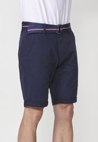 Homme portant un short bleu marine décontracté avec des ourlets retroussés et une ceinture en tissu bleue avec des rayures rouges et blanches, associé à un t-shirt blanc.