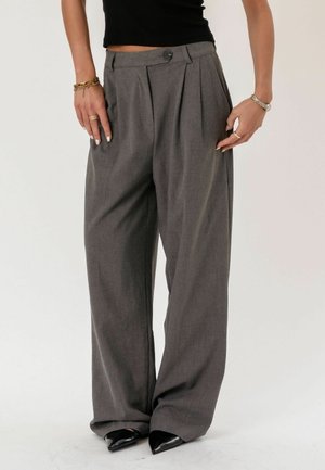 Žena oblečená v sivých nohaviciach s vysokým pásom a širokými nohavicami so záhybmi, doplnených čiernymi špicatými topánkami a zlatými a striebornými náramkami.