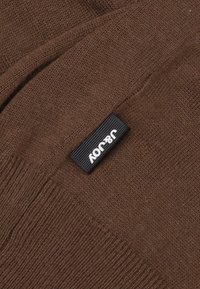 Pull en maille marron au toucher doux, avec une étiquette noire portant un texte blanc. Le design est simple et sans ornements, mettant en avant le matériau.
