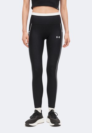 Kobieta nosząca czarne legginsy z wysokim stanem z białym wykończeniem i logo Under Armour, w połączeniu z czarno-białymi butami sportowymi i białymi skarpetkami.