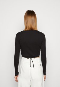 Svart rippad långärmad topp med en kortad design och justerbart knyt bak, i kombination med höga vita byxor.