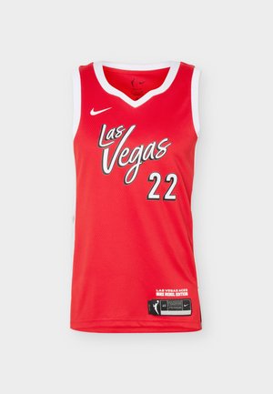 Fiatal férfi sportoló, aki piros Las Vegas Aces kosárlabda mezben, a 22-es számmal és fekete sportnadrágban áll fehér háttér előtt.