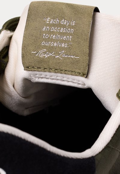 Bato padas su alyvuogių žalios spalvos etikete, ant kurios siuvinėta citata „Kiekviena diena yra proga save iš naujo atrasti.“ pasirašyta Ralph Lauren.