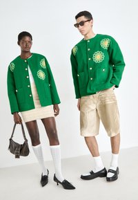 Grønn ulljakke med blomsterbroderi, beige teksturert skjørt, gullshorts, sorte spisse sko, hvite knelange sokker og solbriller.