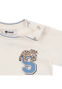 Chemise pour bébé de couleur crème avec col à bordure bleue, boutons-pression, et un léopard reposant sur une grande lettre "S" bleue imprimée sur le devant.