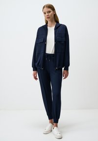 Un survêtement de couleur marine composé d'une veste à capuche avec deux poches avant et des poignets élastiques, associé à un pantalon de jogging assorti et des baskets blanches.