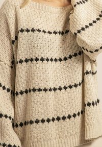 Pull en tricot crème avec des rayures noires horizontales et des motifs d'étoiles. Coupe décontractée avec des manches larges et un col côtelé.