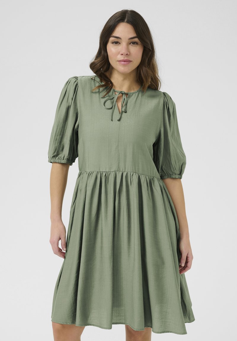 Grünes, knielanges Kleid aus leichtem Stoff. Verfügt über Puffärmel, einen gebundenen Ausschnitt und eine geraffte Taille mit einem fließenden Rock.