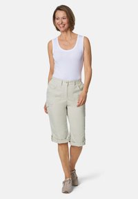 Lichtbeige cargobroek met opgerolde pijpen, voorzien van zijzakken en een knoopsluiting, gecombineerd met een witte tanktop en grijze sneakers.
