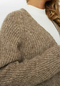 Texturerad beige cardigan med en lös stickad design, breda ärmar och en djup V-hals, lager på lager över en vit ribbad polotröja.