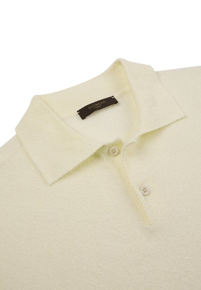 Kremowa teksturowana koszulka polo z dwoma guzikami i miękkim kołnierzykiem, z czarną etykietą z złotym napisem.