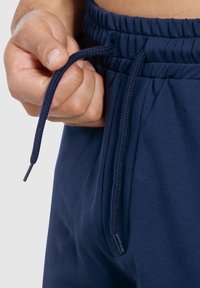 Pantalones de chándal azul marino con cintura ajustable mediante cordón, tejido texturizado y una mano sosteniendo el cordón, mostrando una vista cercana de la zona de la cintura.
