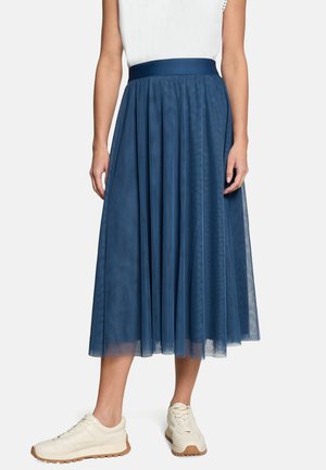 Frau trägt einen marineblauen Midi-Tüllrock mit hohem Bund, weißem Oberteil und weißen Sneakers, steht vor einem einfarbigen Hintergrund.