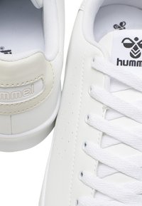 Baskets blanches avec une tige en cuir lisse, des détails perforés, des lacets blancs plats et une languette en tissu arborant le logo Hummel.