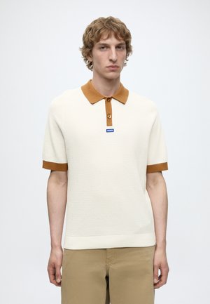 Jeune homme aux cheveux blonds bouclés portant un polo en maille blanc à manches courtes avec un col et des poignets marron, ainsi qu'un pantalon beige, regardant droit devant.
