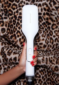 Main avec vernis à ongles rouge tenant une brosse lissante ghd duet style blanche contre un fond en tissu léopard.