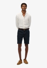 Camisa de lino blanca de botones, pantalones cortos azul marino, cinturón de cuero marrón y zapatos casuales color arena. El modelo está de pie contra un fondo liso.