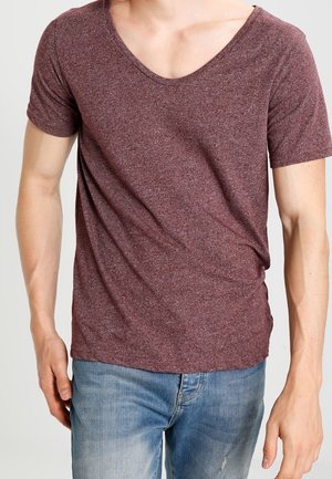 Hombre con camiseta de manga corta en color burdeos y cuello en V, y jeans azul claro, de pie con las manos relajadas a los lados.