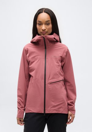 Frau trägt eine staubrosafarbene Jacke mit hohem Kragen und schwarzem Reißverschluss, steht vor einem schlichten weißen Hintergrund.