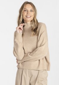 Beżowy sweter z golfem w miękkim, teksturowanym dzianinie z prążkowanymi mankietami i dołem, o luźnym kroju i wyraźnymi bocznymi rozcięciami.